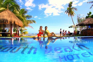 Una piscina con la scritta 'South Beach Hotel' sulla superficie, circondata da palme e ombrelloni di paglia.
