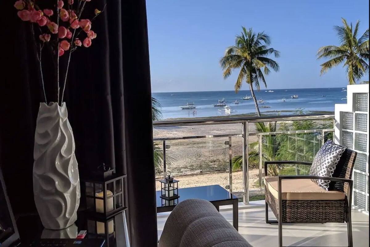 Un balcón con vista a la playa y al océano, una silla de mimbre y un florero con flores.