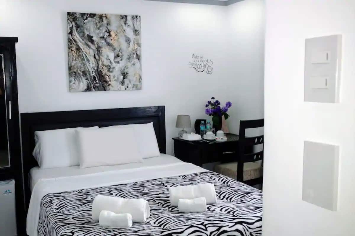 Una habitación de hotel con una colcha de estampado de cebra, un cuadro abstracto de gran tamaño, un interruptor de luz en la pared y una pequeña mesa con un jarrón de flores.