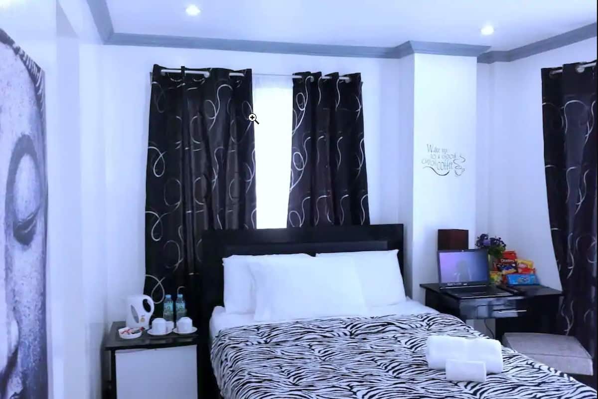 Una habitación de hotel con una cama, una mesita de noche con una computadora portátil, una pequeña mesa con aperitivos y una pared con una cita motivacional.