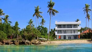 Ein modernes Strandhaus mit zwei Etagen, blauem Wasser, Palmen und einem Sandstrand.