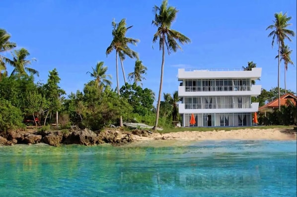 En modern villa i två våningar som ligger direkt vid stranden, med kristallklart blått vatten, palmer och en sandstrand.