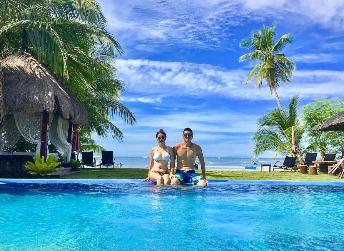 A couple sitting by a pool with a view of the ocean and palm trees.