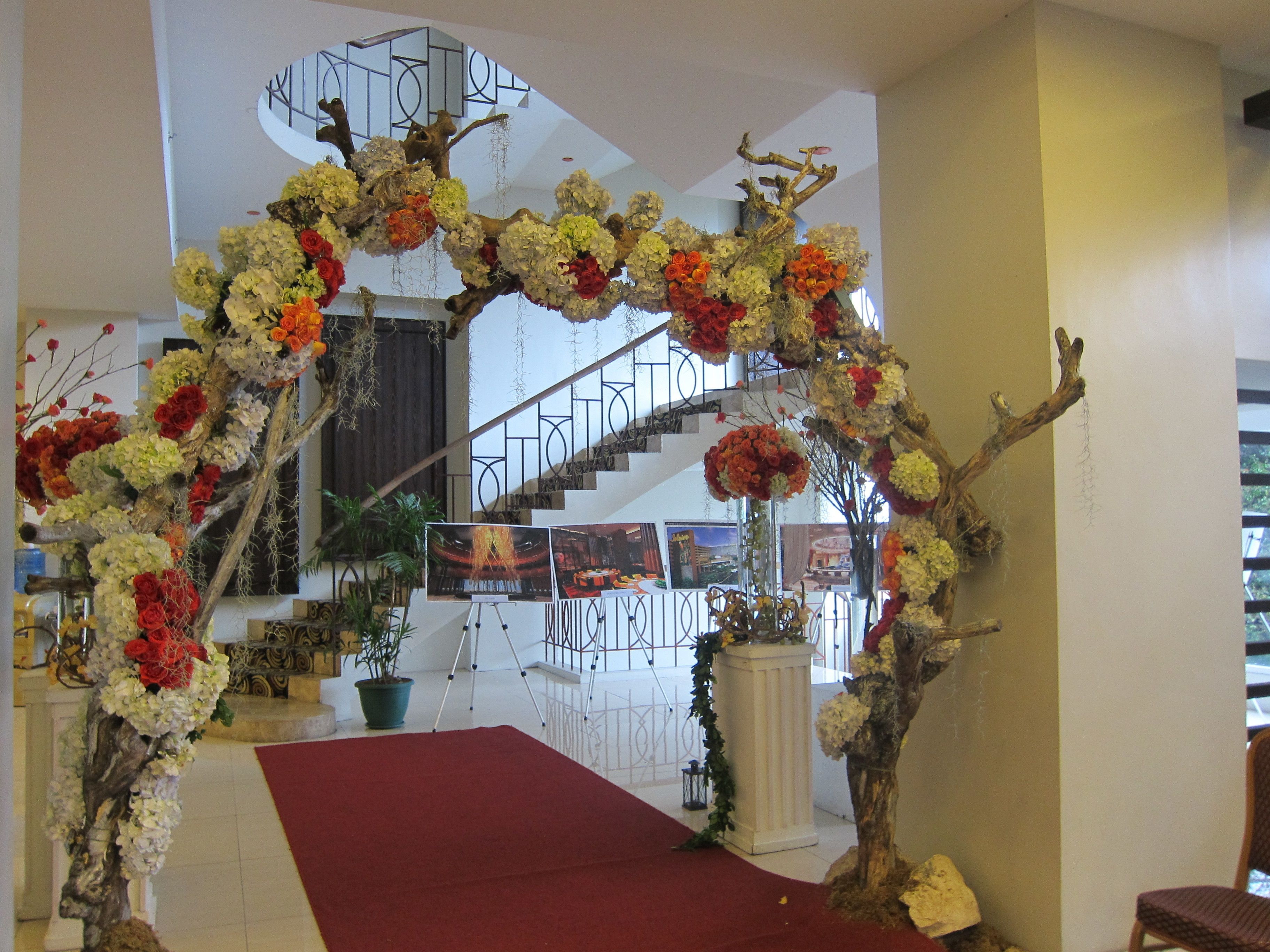 Una escalera con alfombra roja que asciende, rodeada de adornos florales y un columnario blanco.