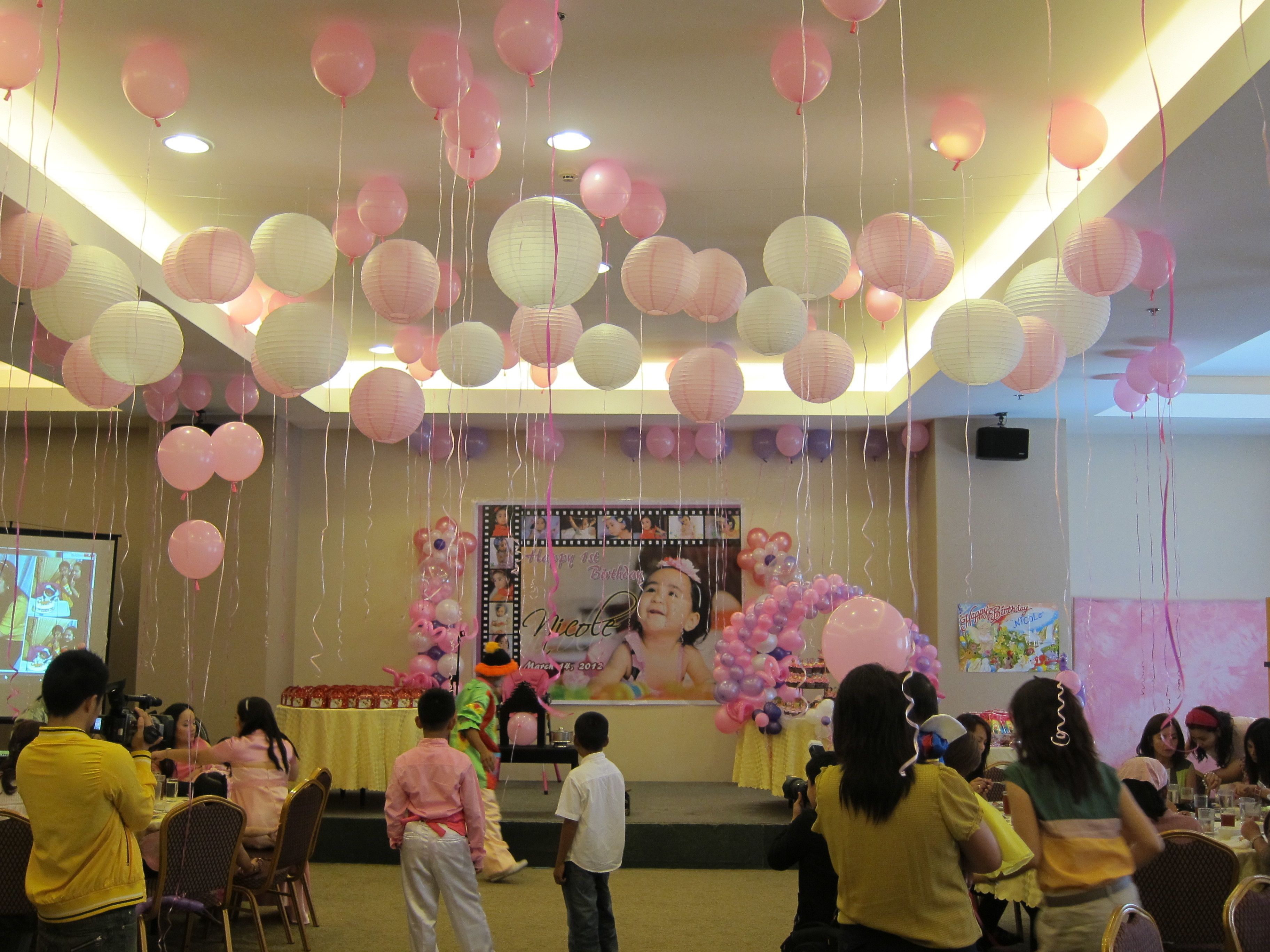 A room with a stage decorated with balloons and paper lanterns, a projector screen, and people seated at tables.