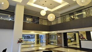 Un hall d’hôtel moderne avec un plafond haut, de grands luminaires décoratifs, une balustrade en verre à l’étage supérieur et un comptoir de réception agrémenté d’un arrangement floral.