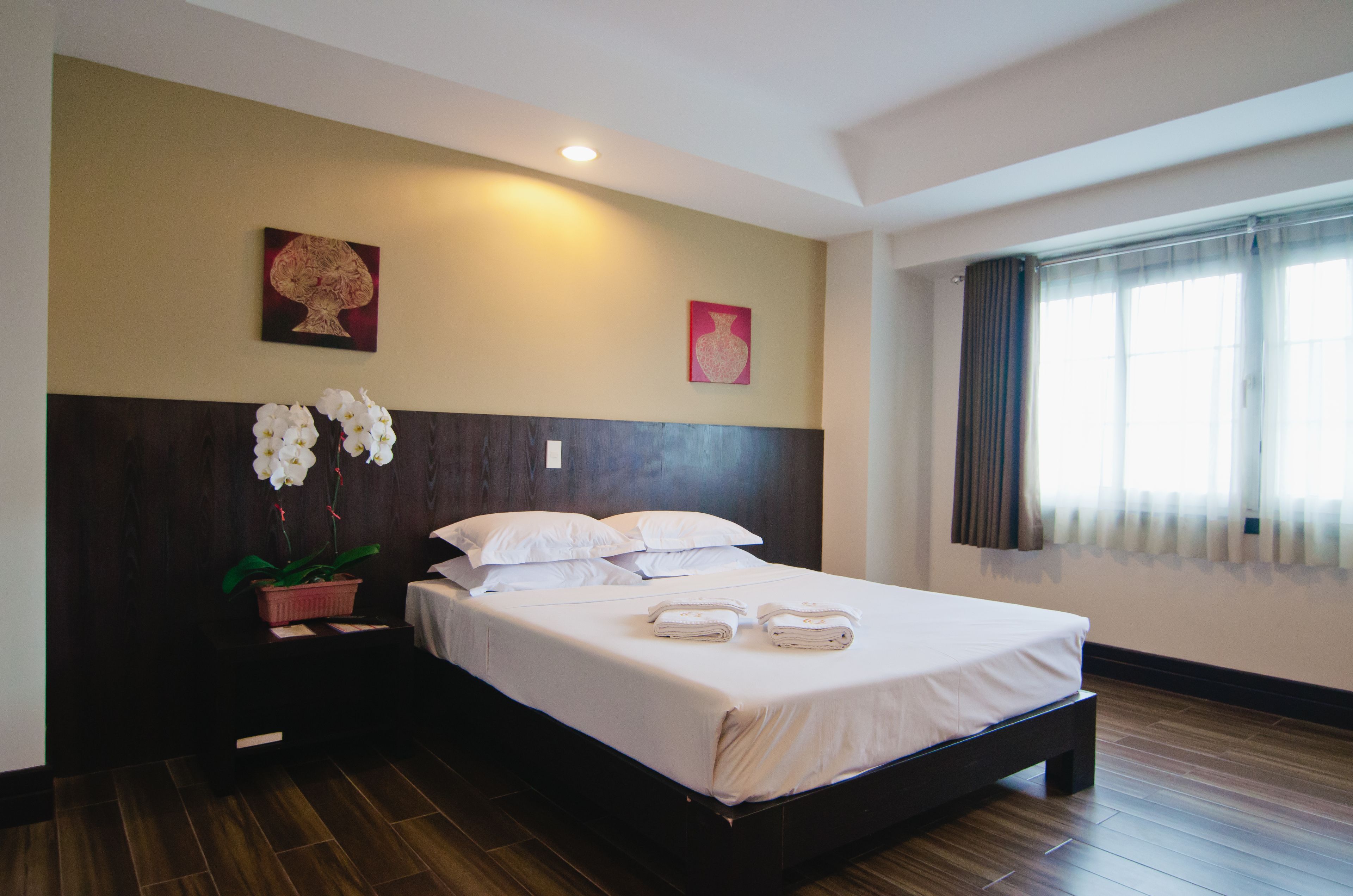 Una cama bien tendida con sábanas blancas, un cabecero de madera y una orquídea en maceta sobre una mesita de noche.