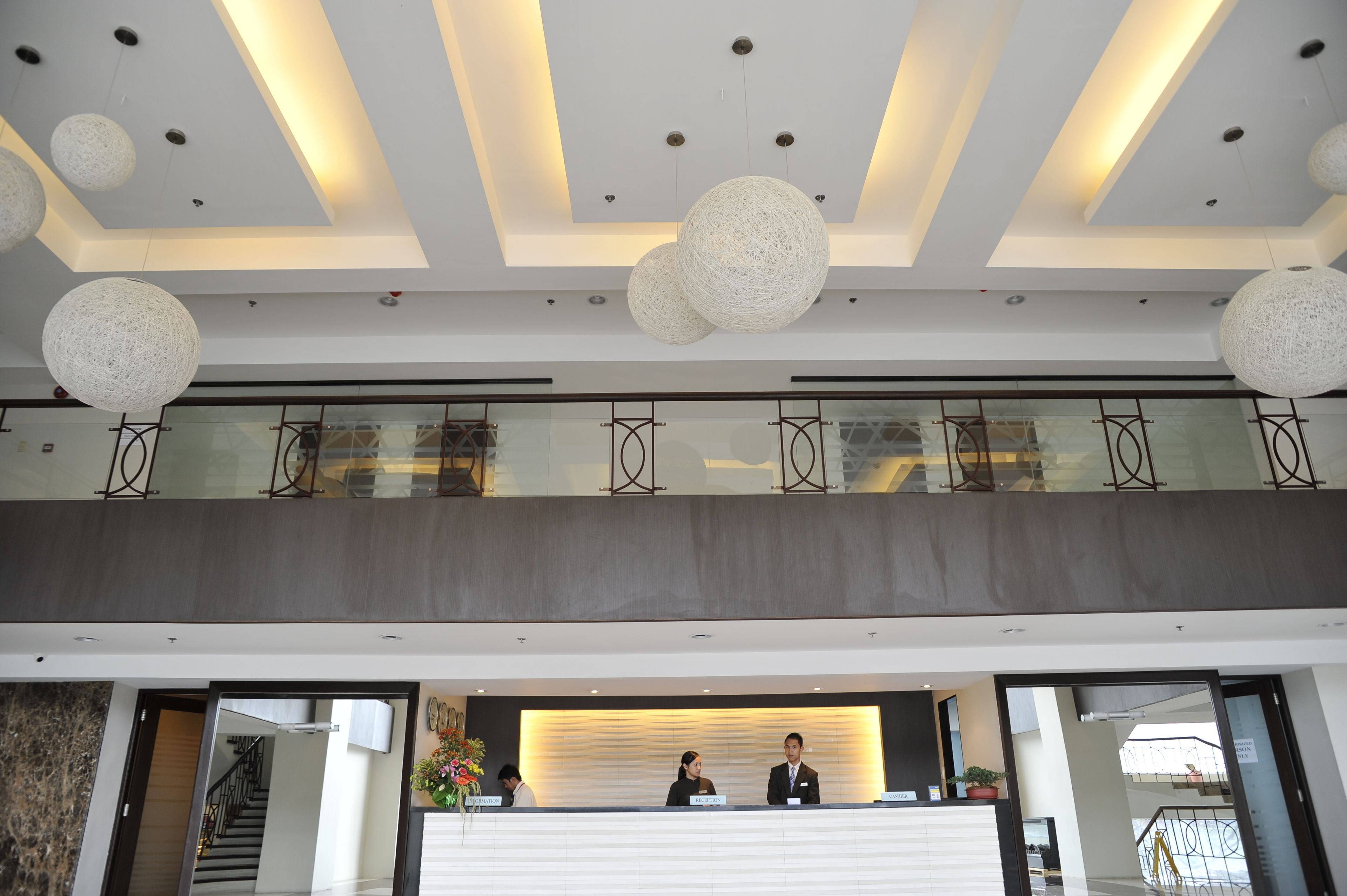 Zona de recepción de hotel con diseño moderno, que incluye un mostrador de recepción, dos empleados y una barandilla de vidrio que da a un nivel inferior.