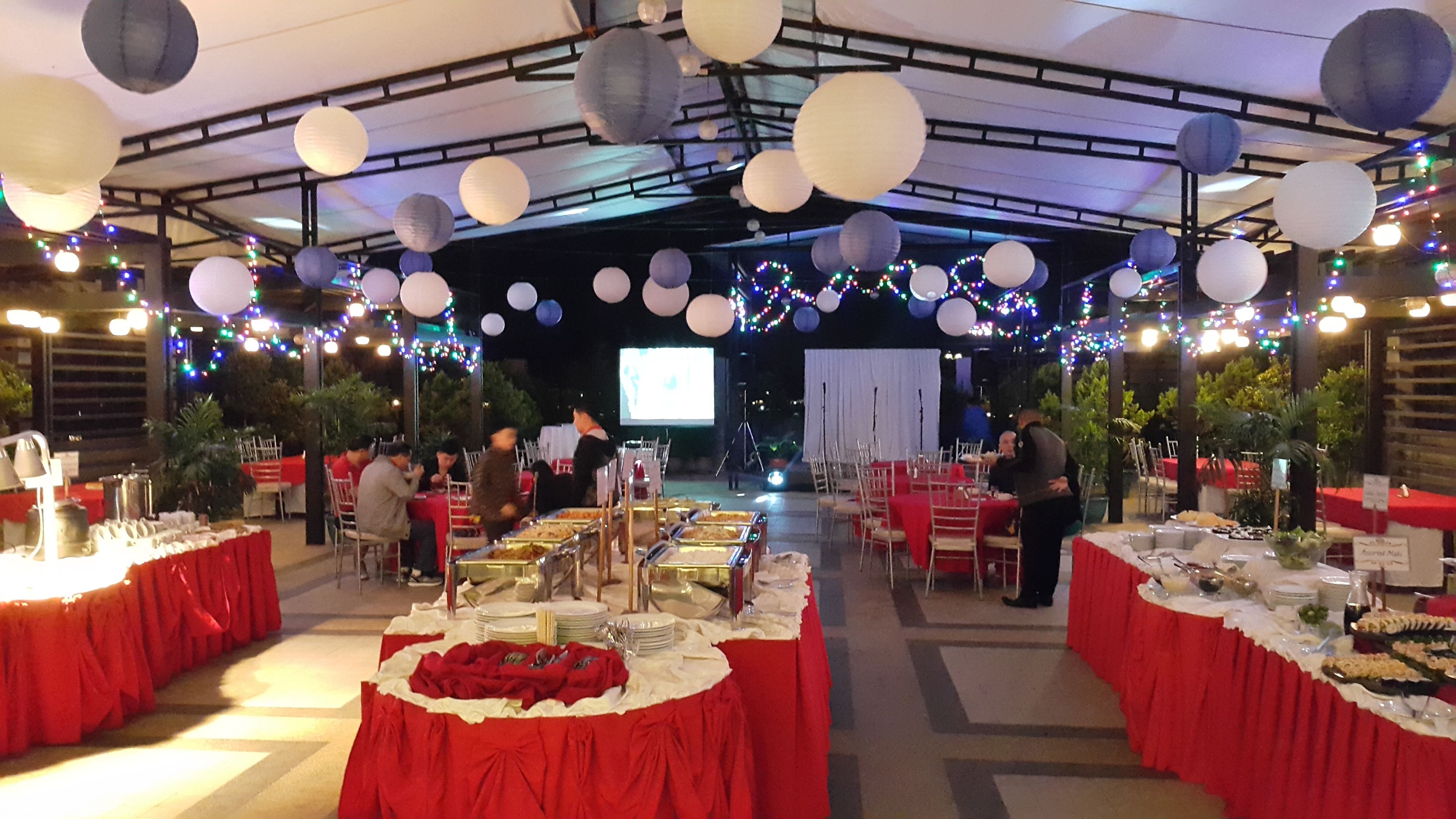 Un salón de banquetes interior con mesas dispuestas para un evento formal, decorado con farolillos de papel colgantes y luces de guirnalda.
