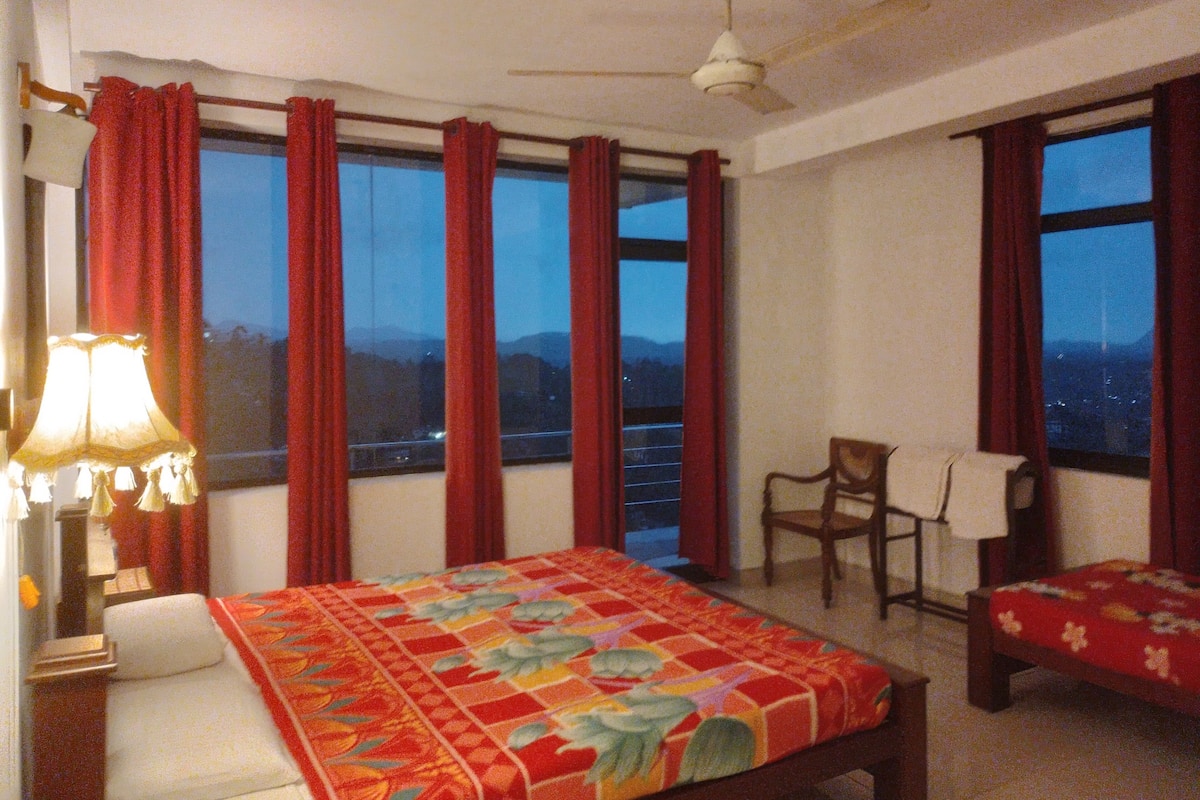A hotel room with a bed, a chair, a desk, and a window with a view of mountains.
