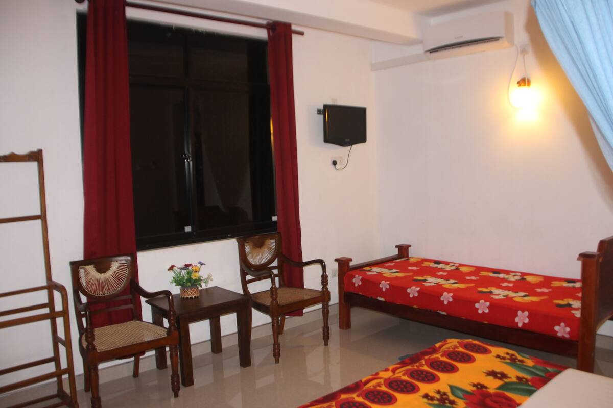 A room with a bed, two wooden chairs, a small table with a vase, a wall-mounted TV, and a window with red curtains.