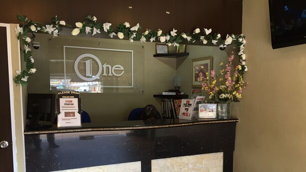 Un mostrador de recepción de hotel con el logo 'One', un punto de entrega de llaves, fotografías enmarcadas y un banco.