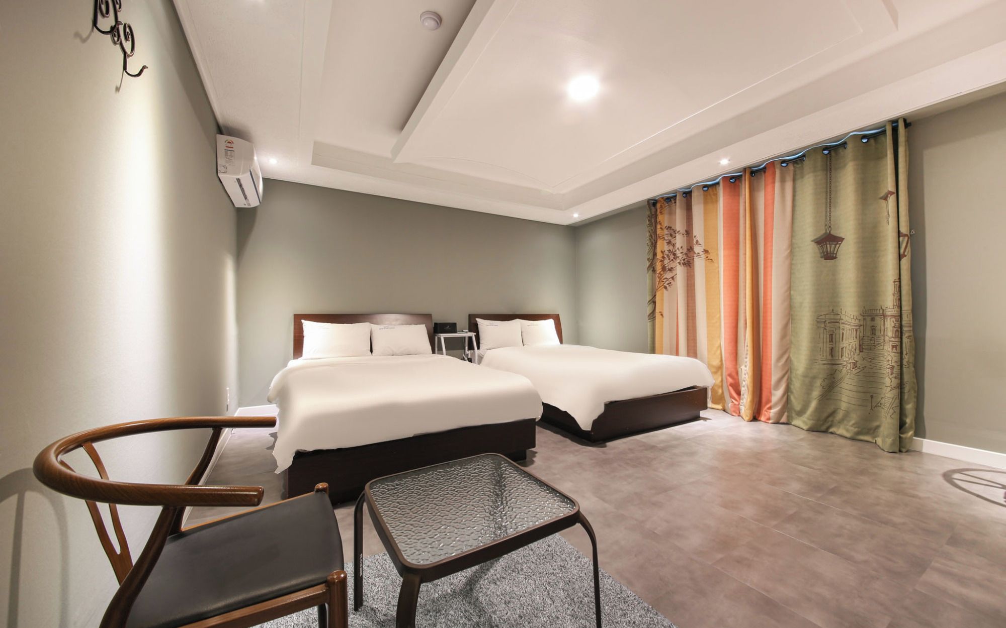 A hotel room with two beds, a small table, a chair, and a curtain with a cityscape design.