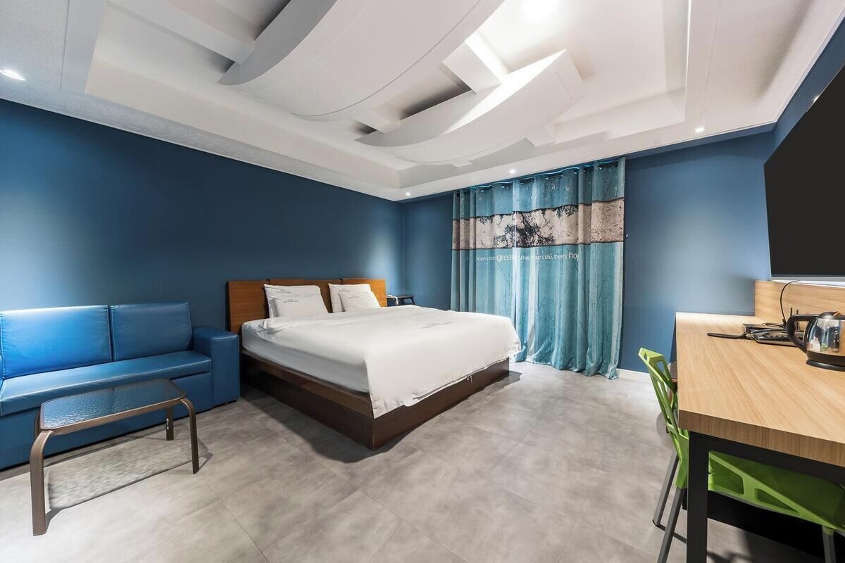 A modern hotel room with a bed, a blue sofa, a wooden desk, and a green chair.