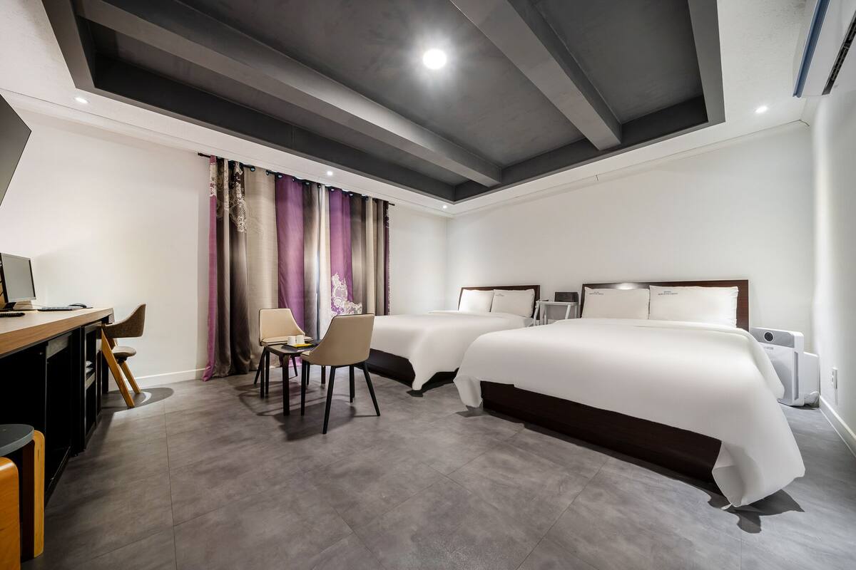 A hotel room with two beds, a desk, chairs, and a large window with curtains.