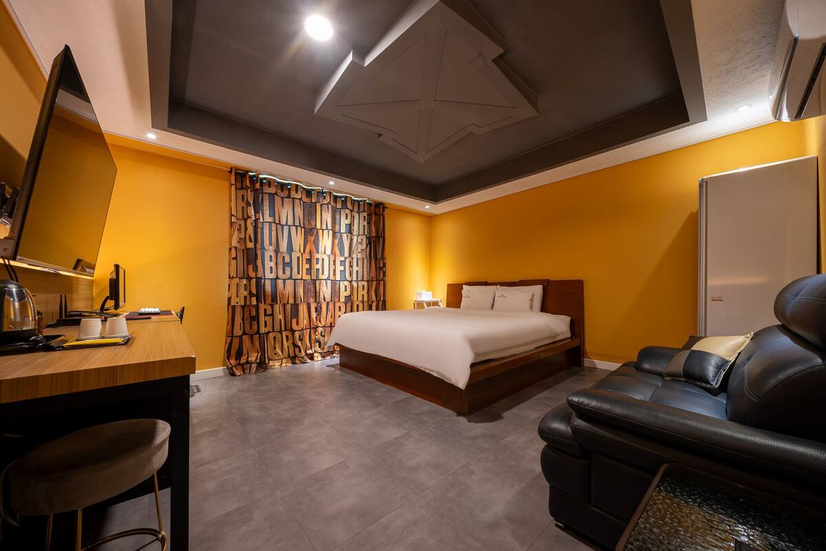 A hotel room with a bed, a desk, a chair, a sofa, a wardrobe, and a mirror.