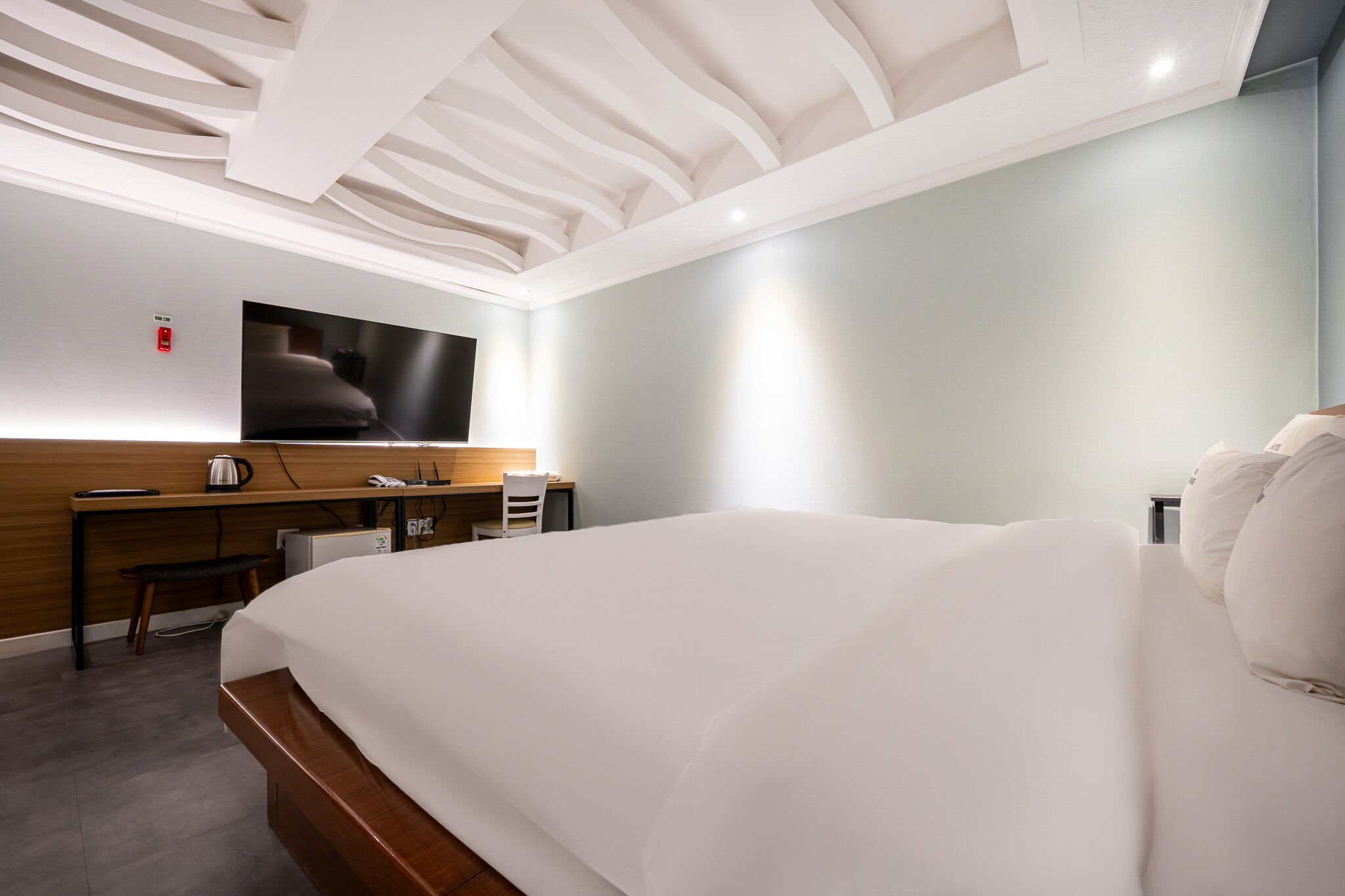A hotel room with a large bed, a desk with a chair, a flat-screen TV mounted on the wall, and a wooden desk with a kettle and a phone.
