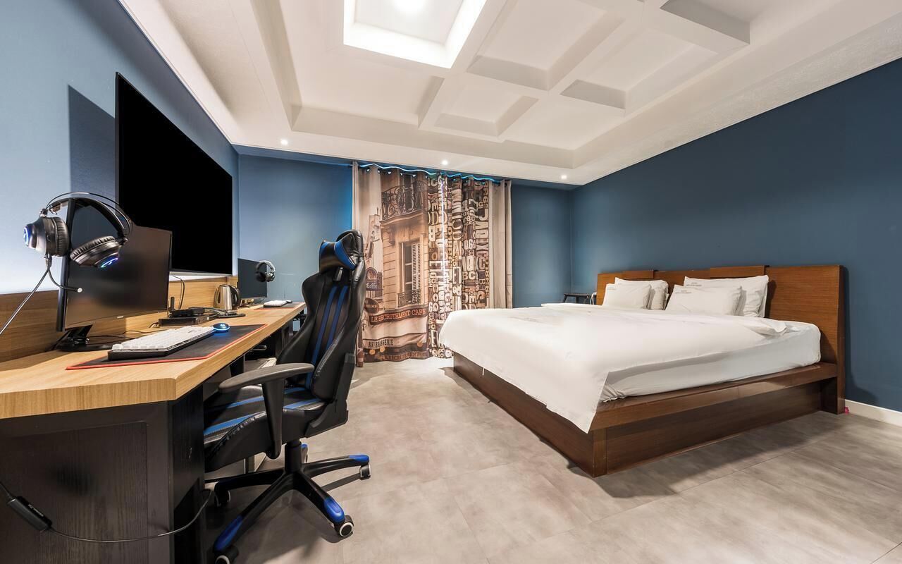 Un dormitorio con escritorio, computadora y auriculares, una cama con almohadas y una cortina estampada.
