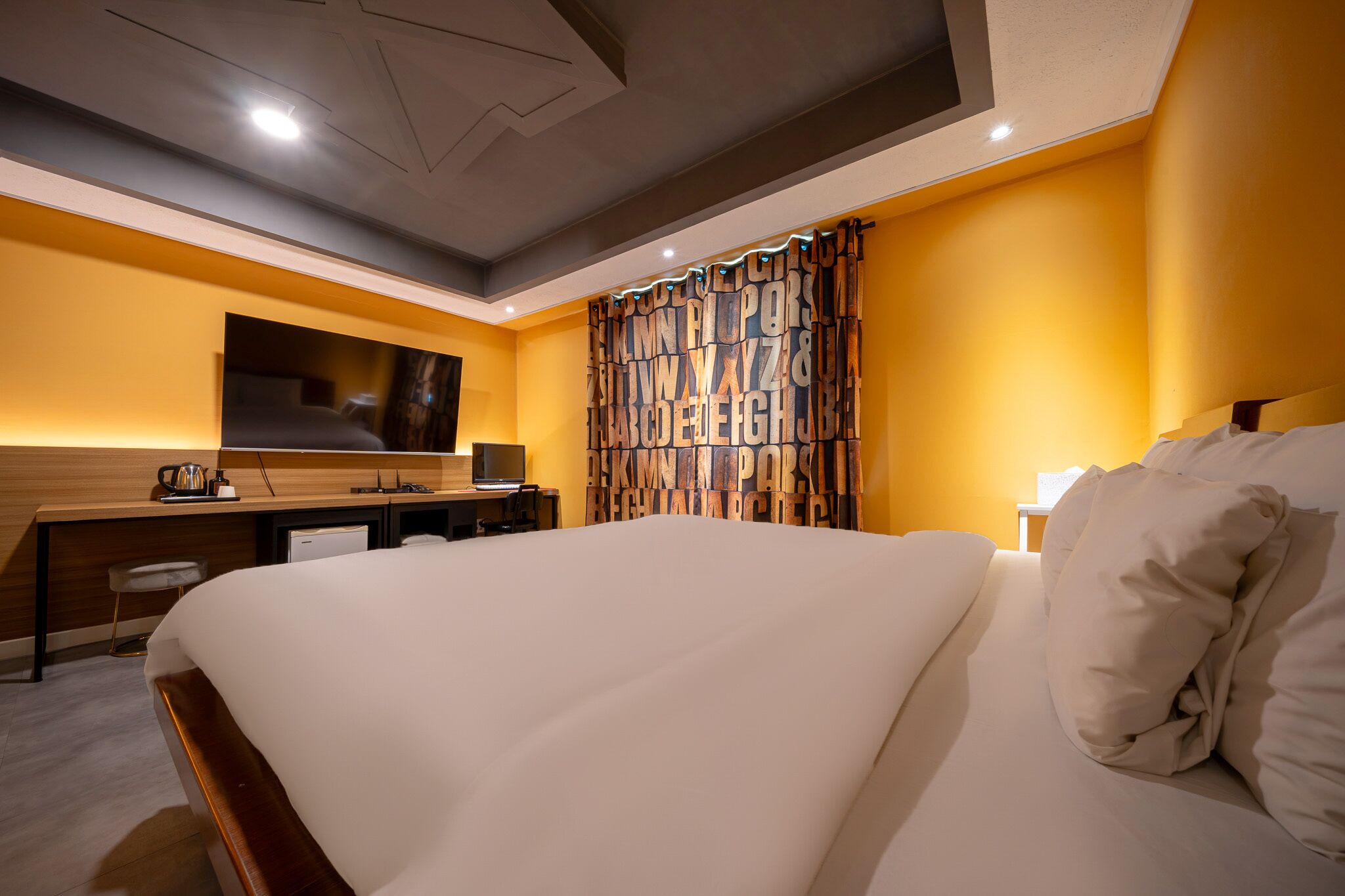 Quarto de hotel com uma cama grande, uma escrivaninha, uma televisão e uma cortina com letras do alfabeto.