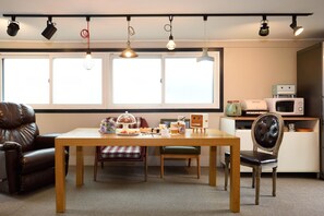 A dining area with a wooden table, chairs, and a buffet setup. There are large windows with blinds, a microwave, and a small refrigerator on the counter. - The One Hostel (Seoul)