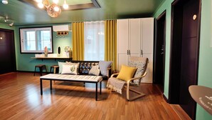 A living room with a sofa, armchair, and coffee table. There is a window with curtains, a ceiling light fixture, and a door. - The One Hostel (Seoul)