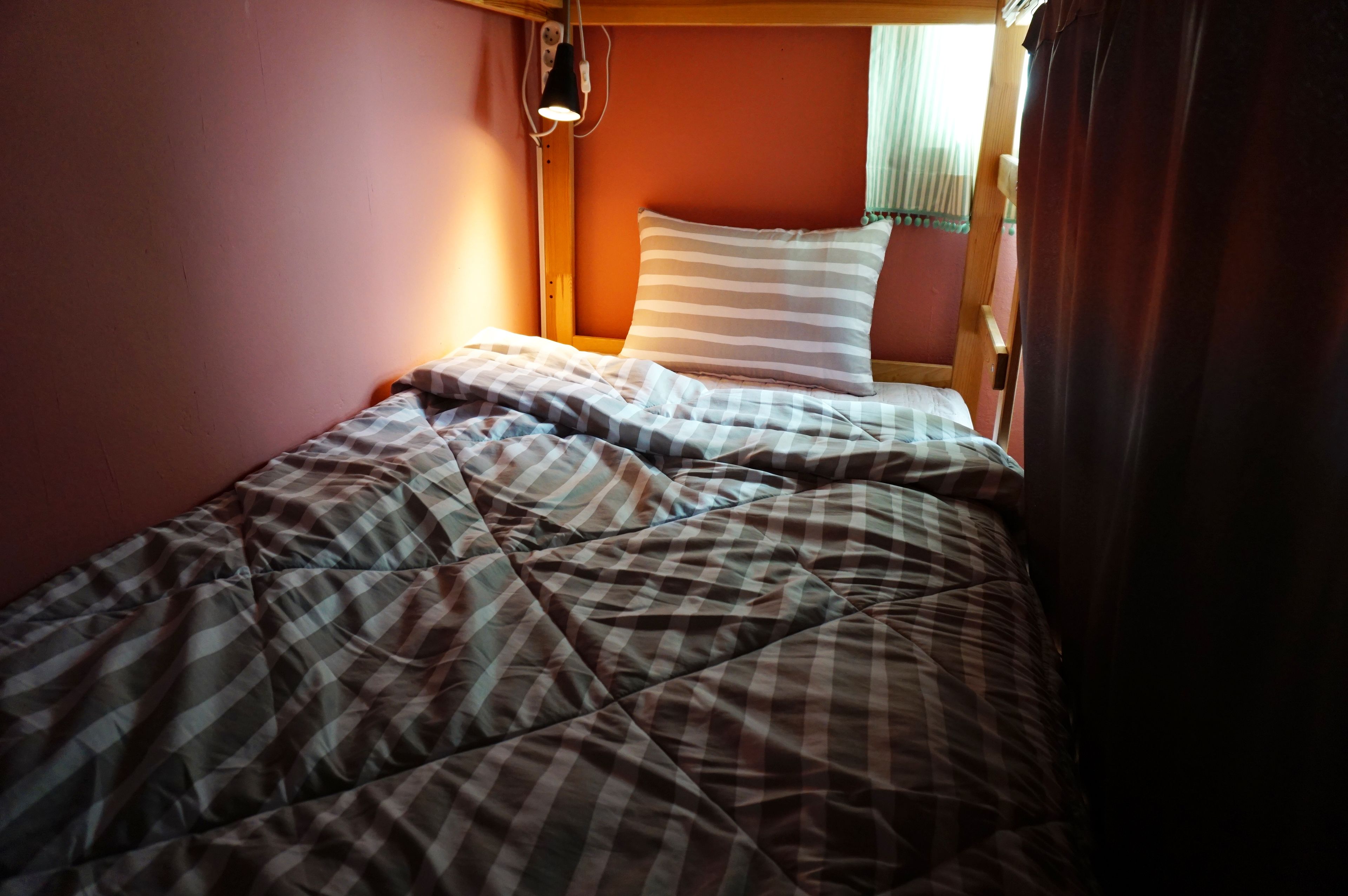 A single bed with a striped pillow and a checkered blanket in a room with a wooden headboard and a wall-mounted lamp.
