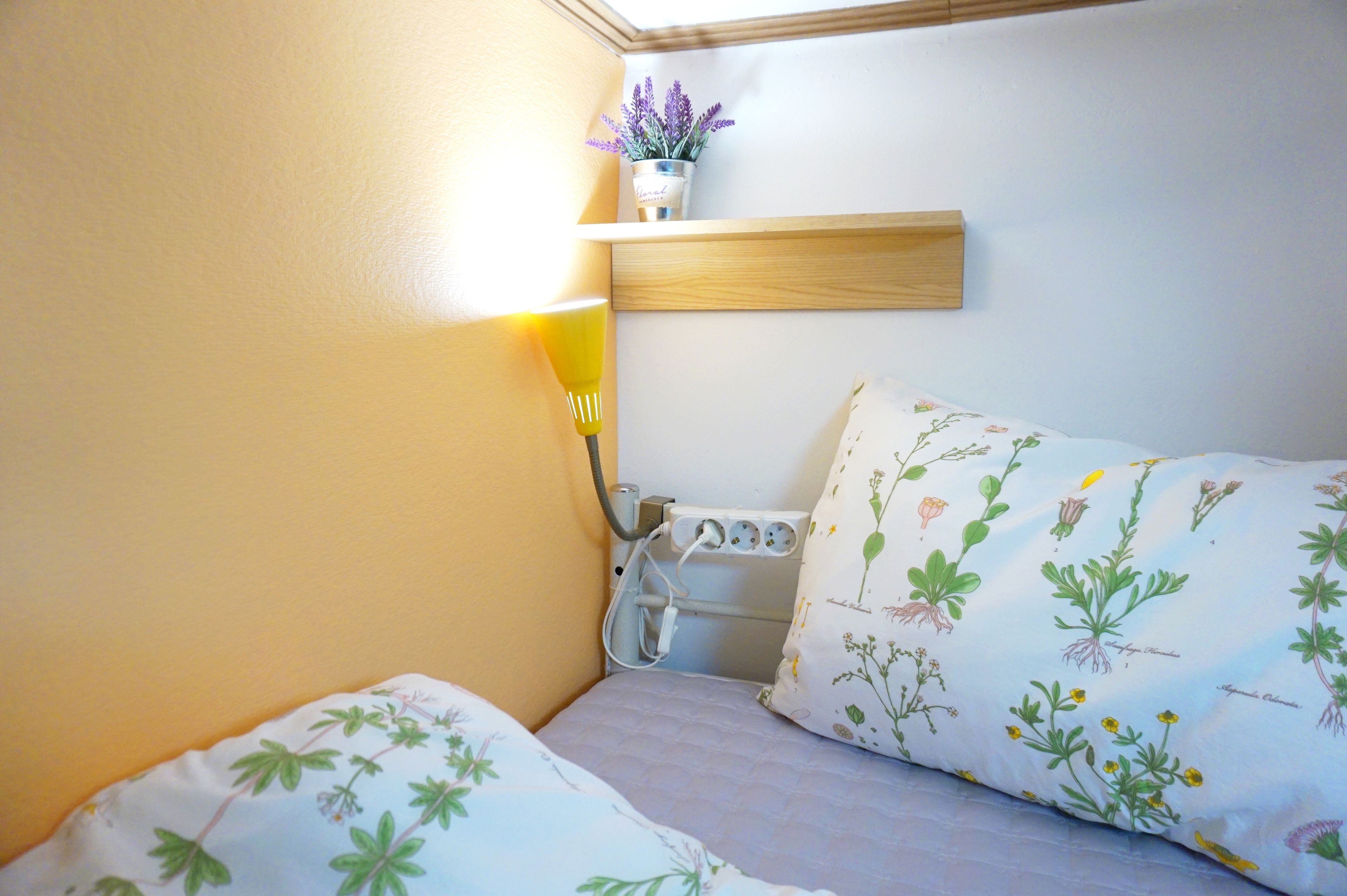 A small, cozy room with a bed, a bedside table, a lamp, and a wall-mounted shelf with a potted plant.