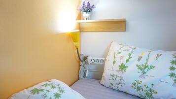 A small, cozy room with a bed, a bedside table, a lamp, and a wall-mounted shelf with a potted plant.
