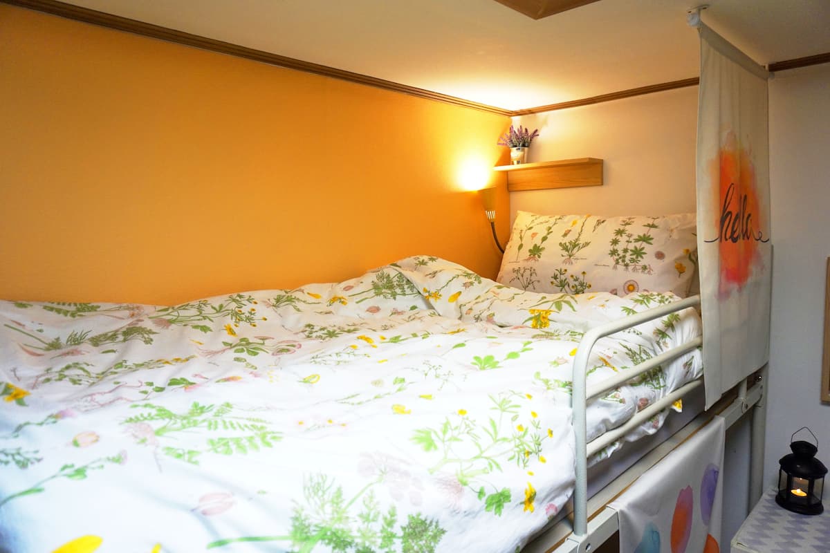 A single bed with floral bedding, a bedside table with a lantern, and a wall-mounted shelf with a plant.