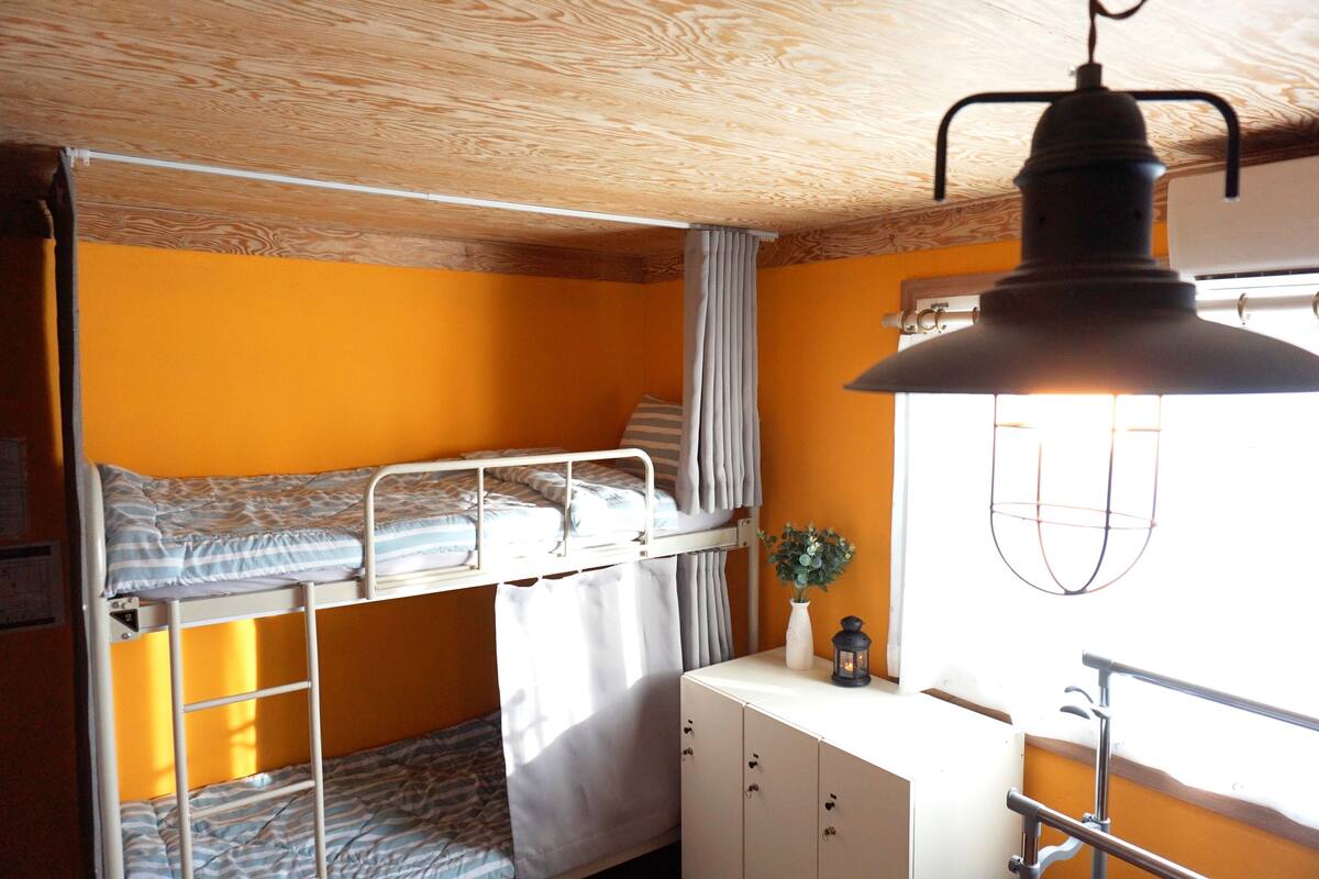 A bunk bed room with a wooden ceiling, a white desk with a vase and a lantern, and a window with curtains.