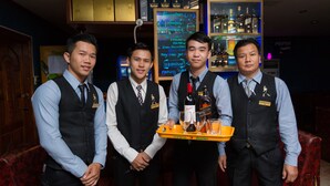 Cuatro hombres en un bar, sosteniendo una bandeja con botellas y vasos.