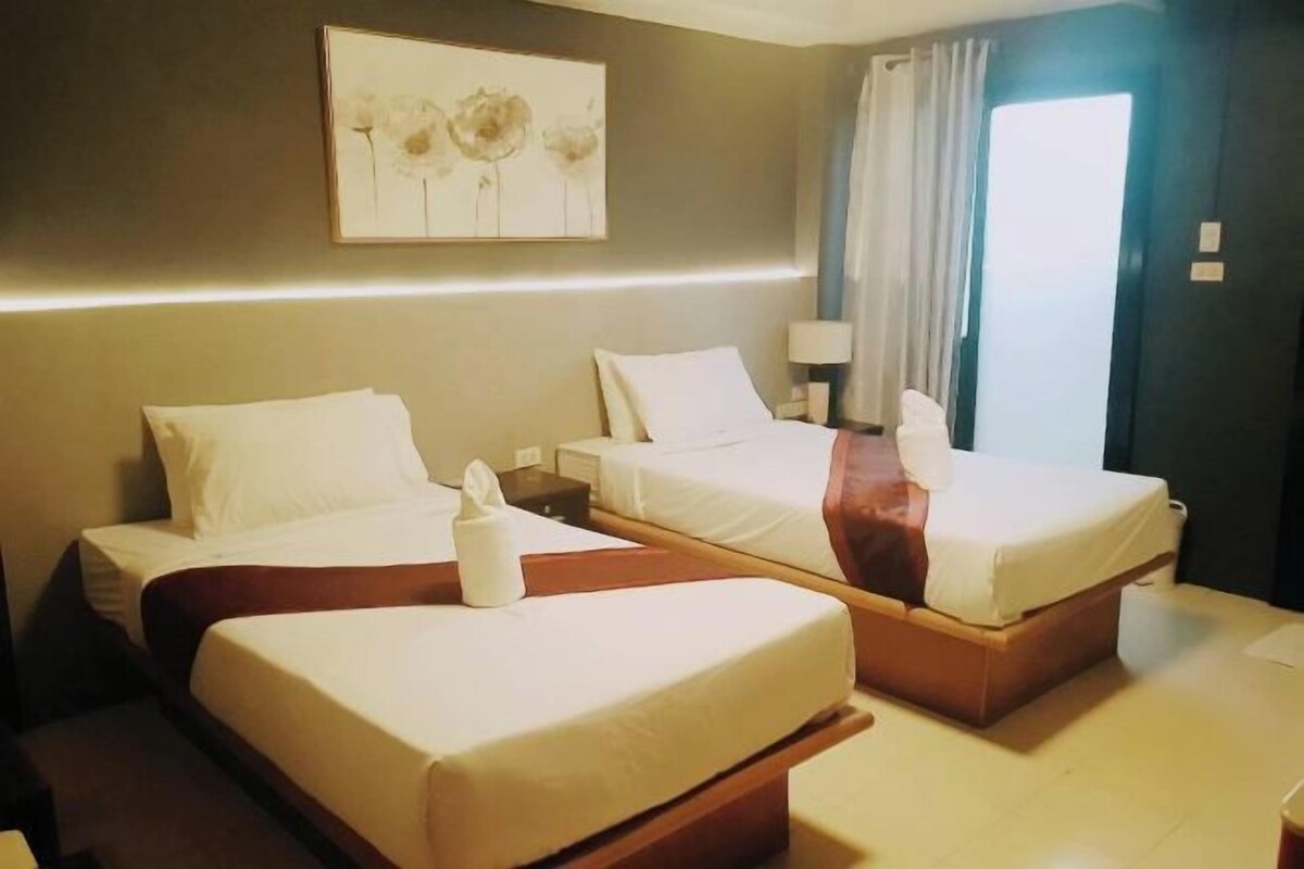 A hotel room with two single beds, a bedside table with a lamp, a painting on the wall, and a window with curtains.