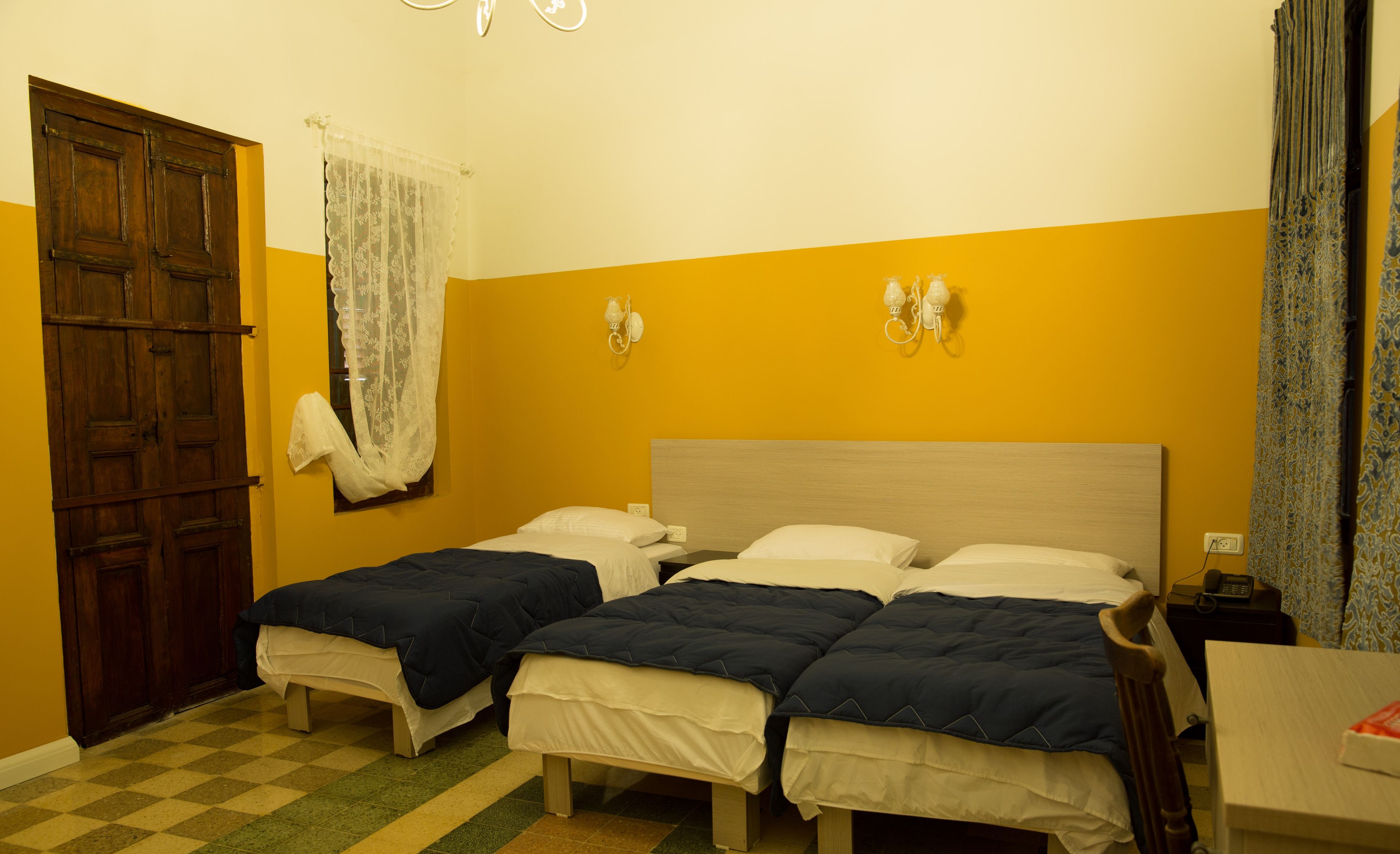 A room with two beds, a wooden door, a checkered floor, and a wall-mounted light fixture.