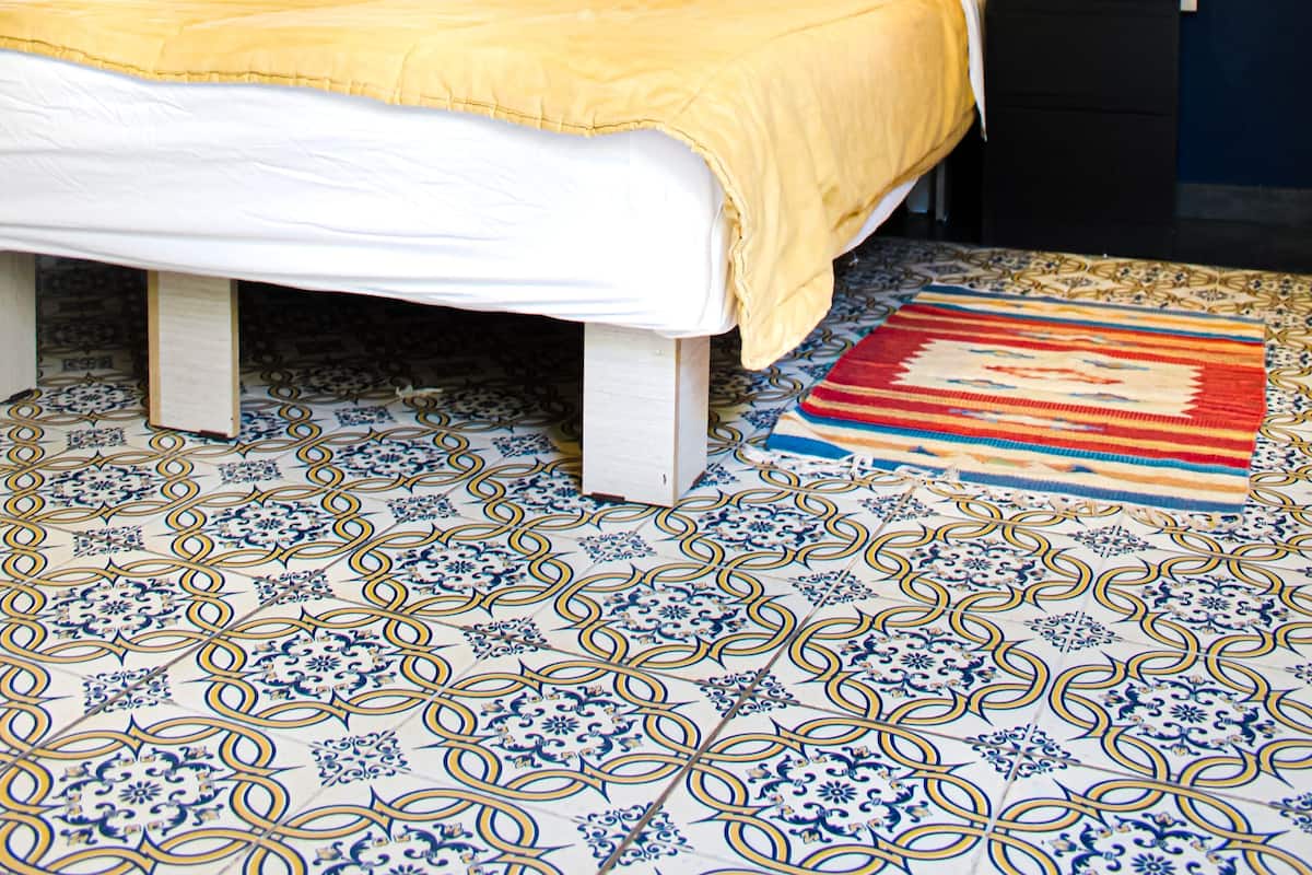Un lit bien fait, recouvert d’une couvre-lit jaune, avec deux oreillers foncés et une table de chevet sur laquelle se trouvent une lampe et un téléphone. La pièce a un mur bleu foncé et un sol recouvert de carreaux à motifs.