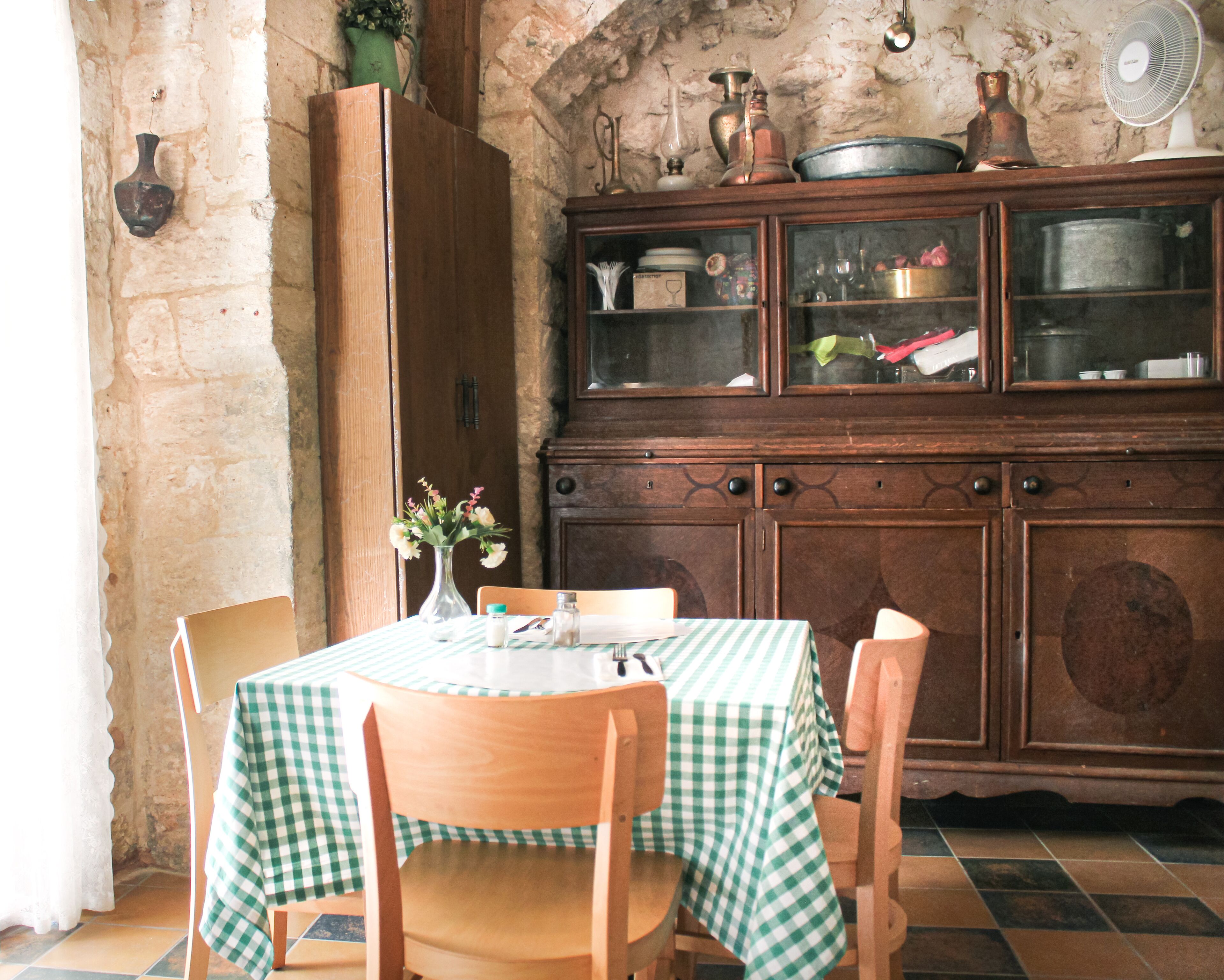 Un comedor con mantel a cuadros, sillas de madera, una alacena con puertas de vidrio y una pared de piedra.