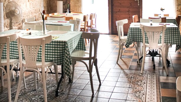 Un coin repas rustique avec des murs en pierre, des poutres en bois et un lustre. Les tables sont dressées avec des nappes à carreaux et des chaises blanches.
