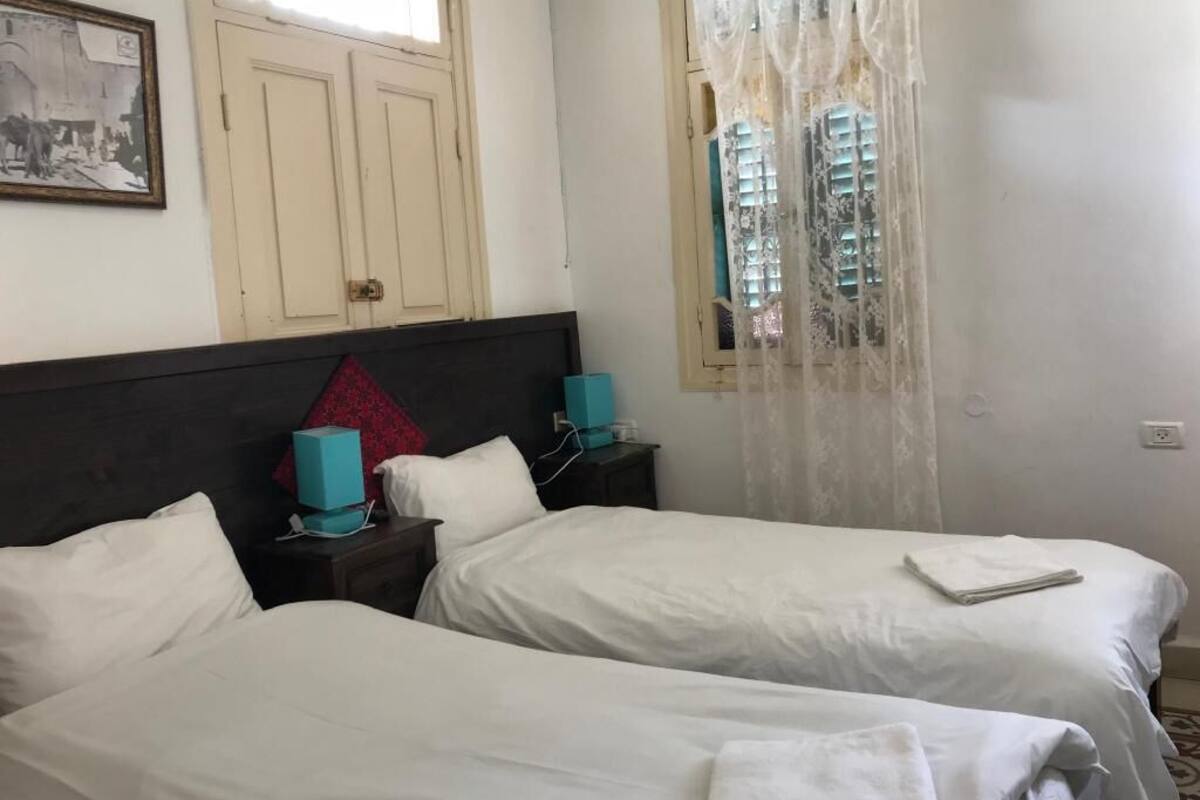 Une chambre avec deux lits simples, une tête de lit en bois, une table de chevet avec une lampe et une fenêtre avec des rideaux.
