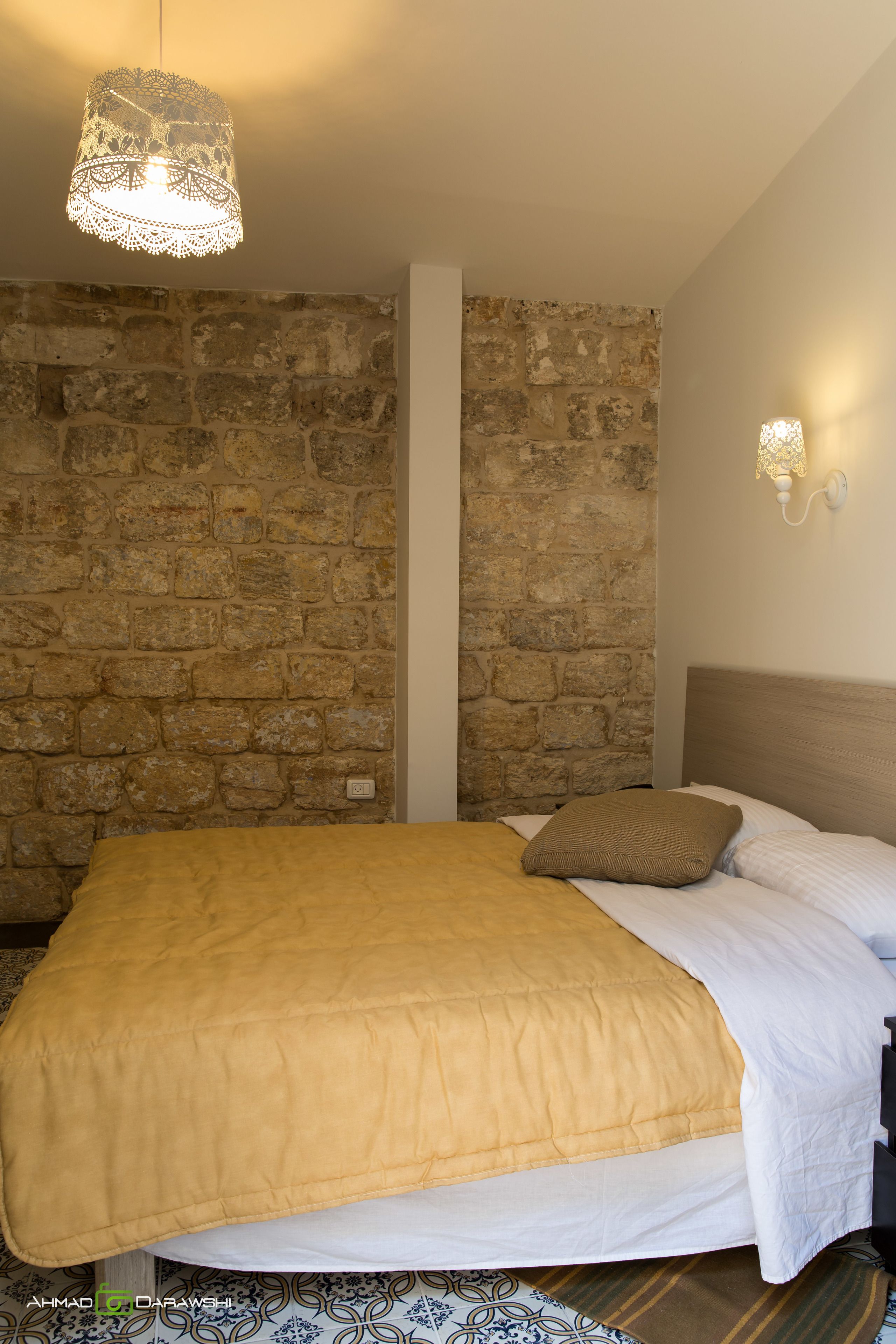A bedroom with a stone wall, a bed with a yellow blanket, a wooden headboard, and a hanging light fixture.