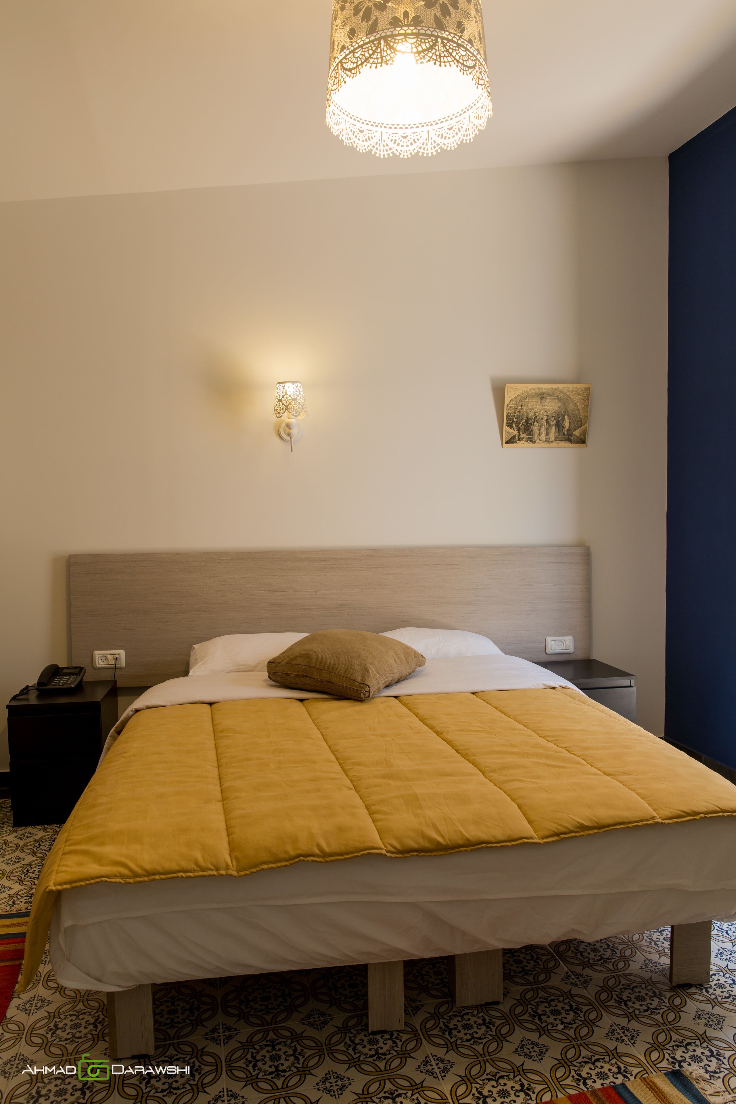 A neatly made bed with a yellow blanket, a bedside lamp, a nightstand with a phone, and a wall-mounted picture.
