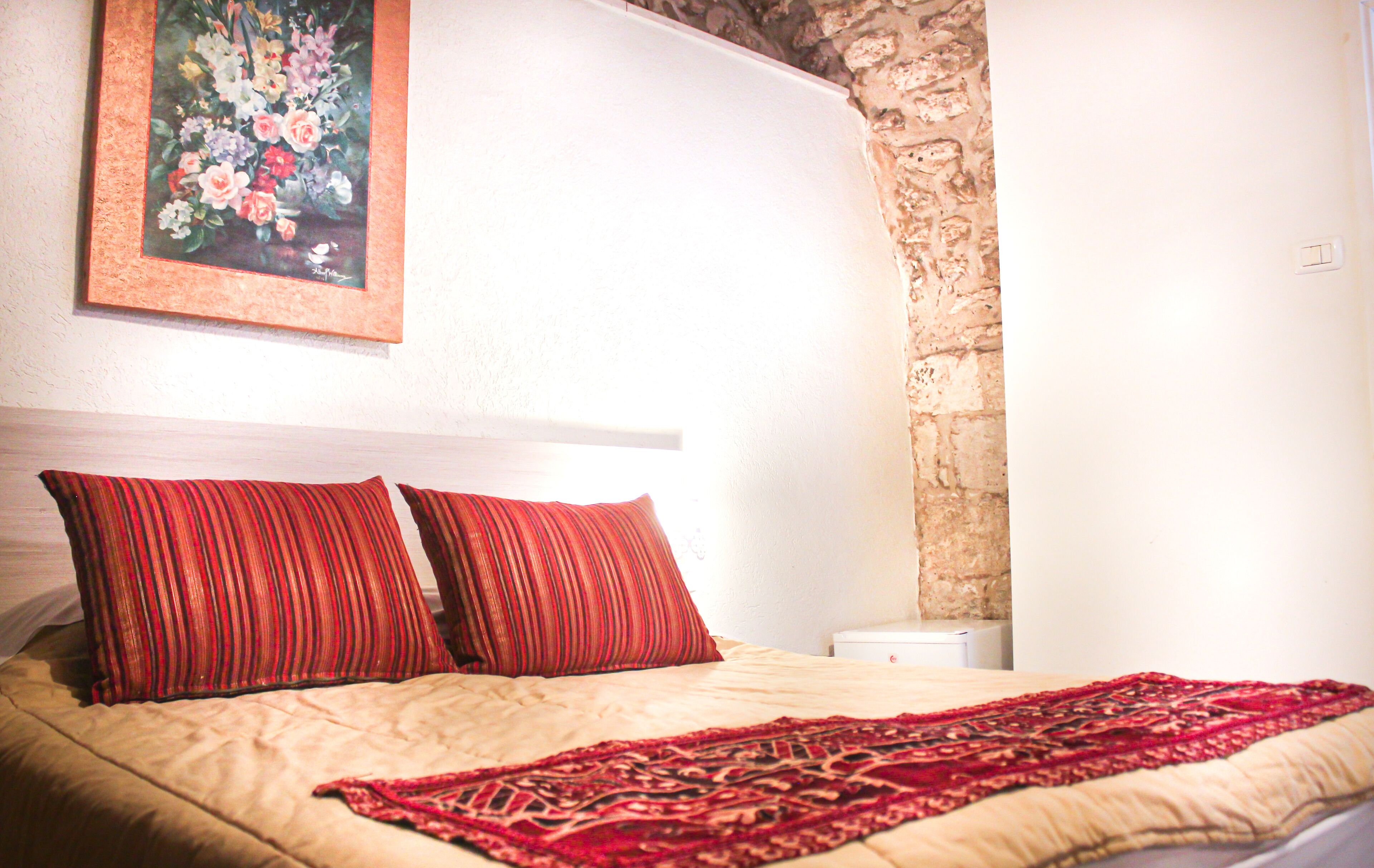 A bed with two red pillows, a red and beige bedspread, and a painting of flowers on the wall.