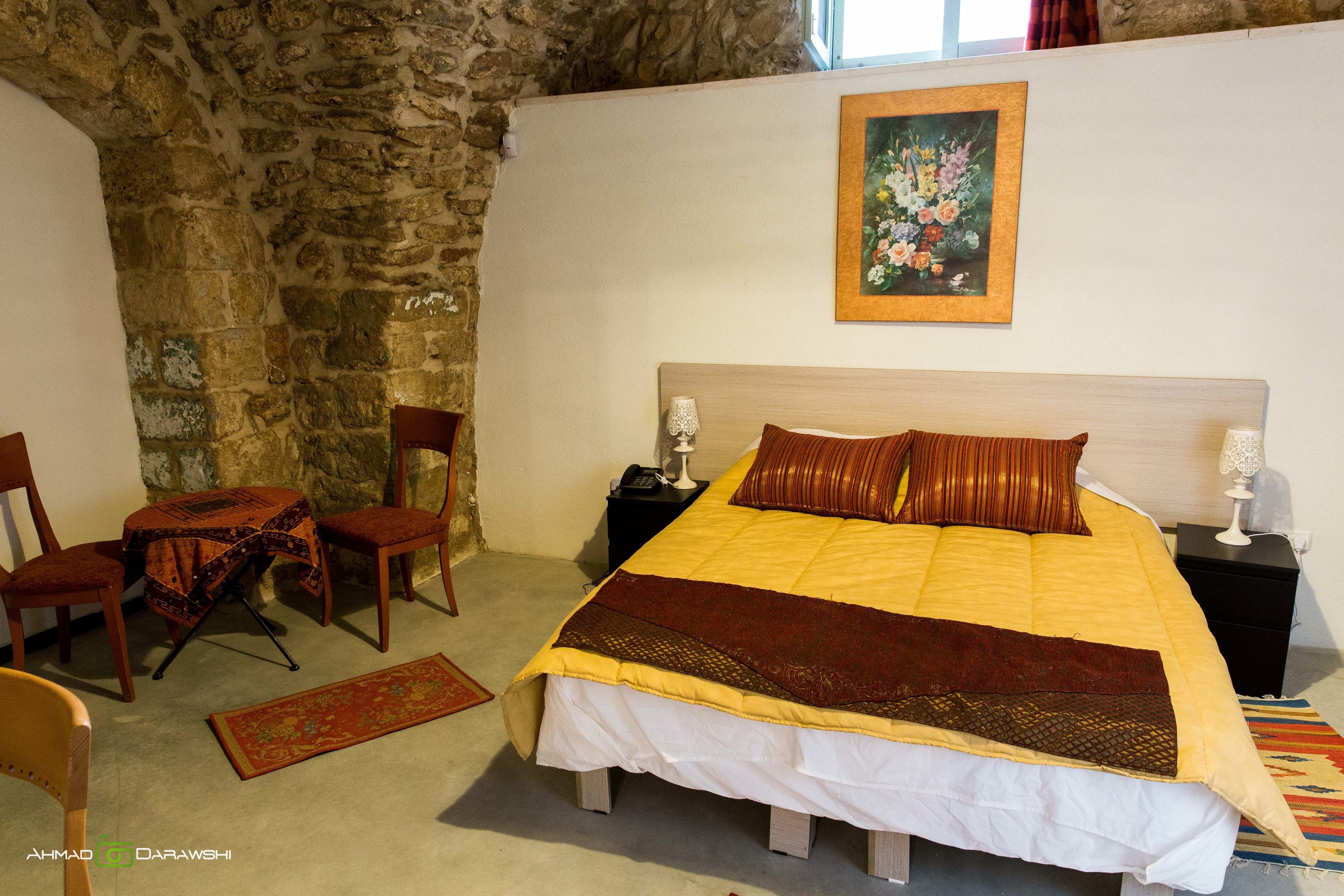 Un dormitorio con pared de piedra, una cama con colcha amarilla y marrón, dos mesitas de noche con lámparas, una silla y una pequeña mesa con mantel estampado.