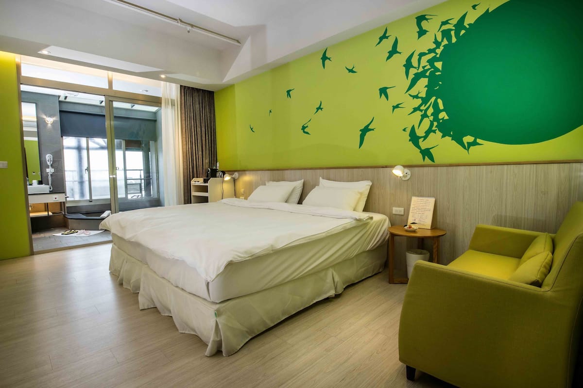 A hotel room with a large bed, a green wall mural, a yellow armchair, and a small table with a lamp.