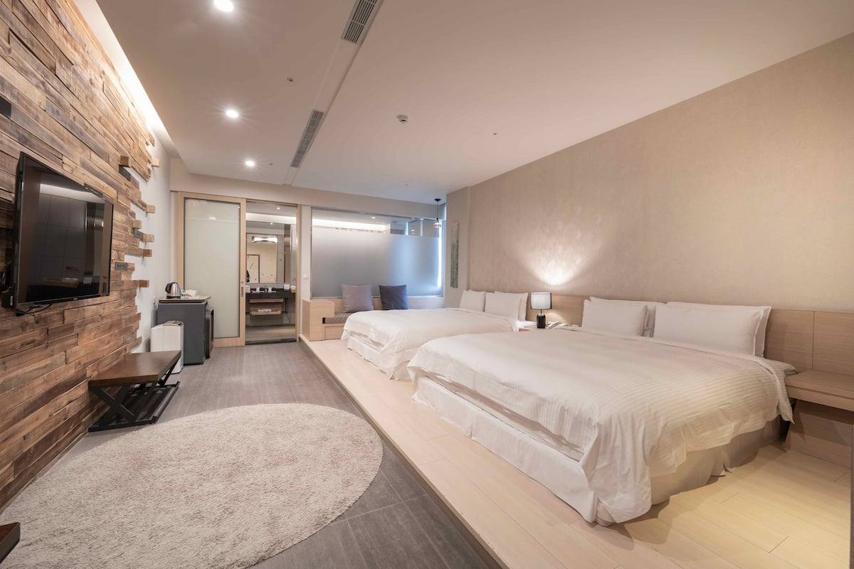 A modern hotel room with two beds, a flat-screen TV mounted on a wooden wall, a desk with a chair, and a large window providing natural light.