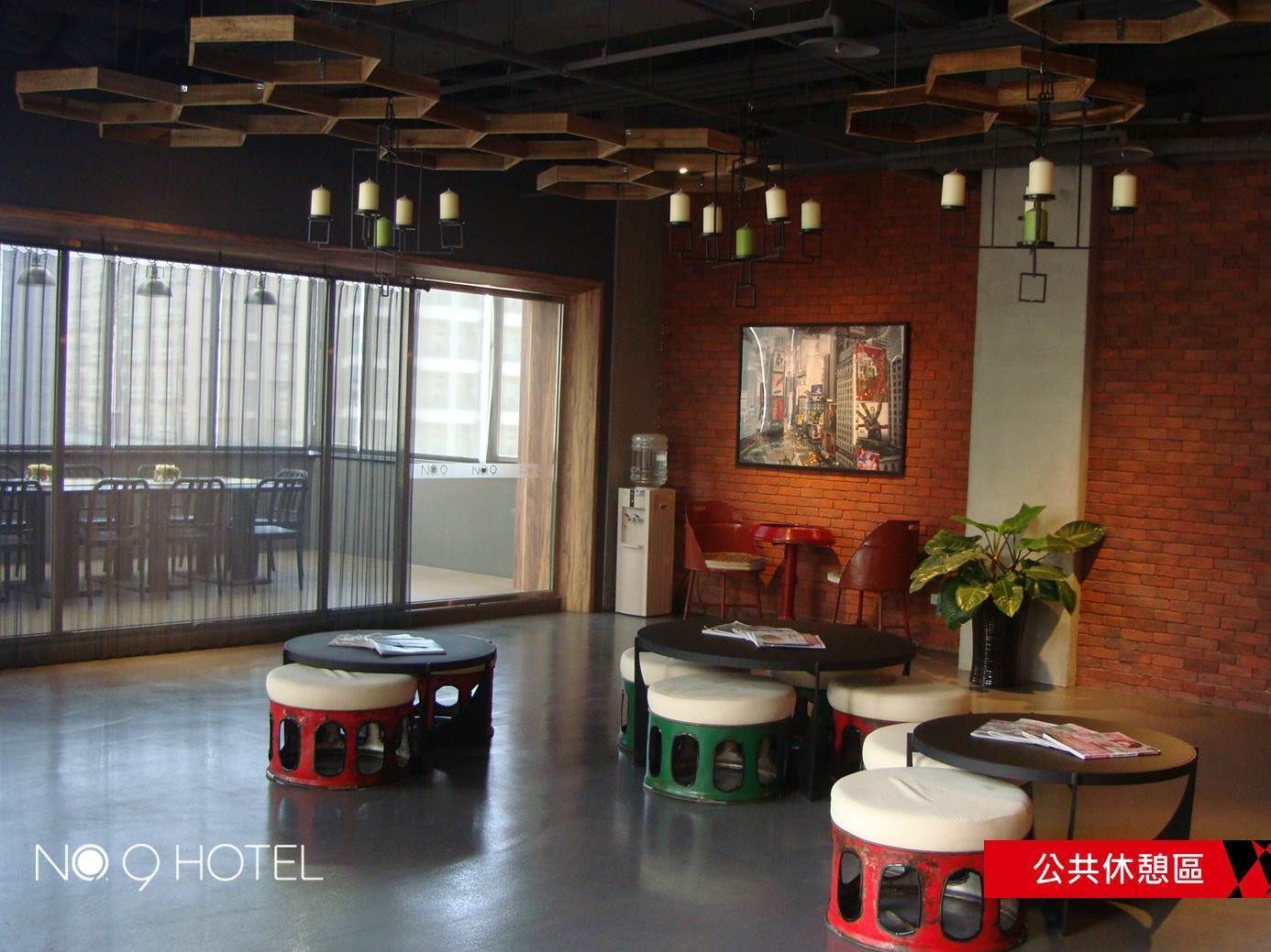 A modern hotel lobby with a large glass door leading to an outdoor area, a brick wall with a cityscape painting, and several round tables with stools.
