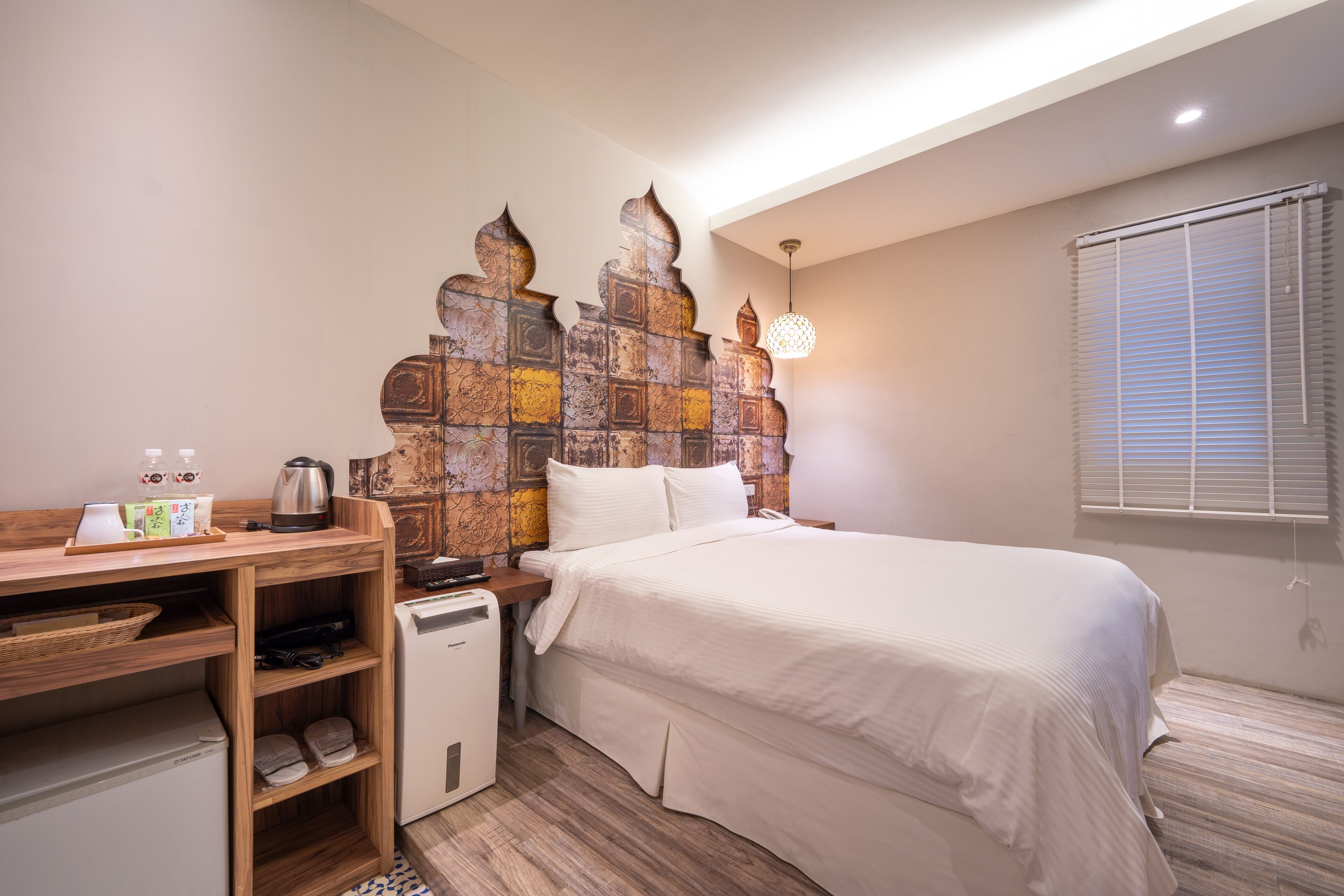 A hotel room with a bed, a desk with a kettle, a small refrigerator, and a decorative wall panel.