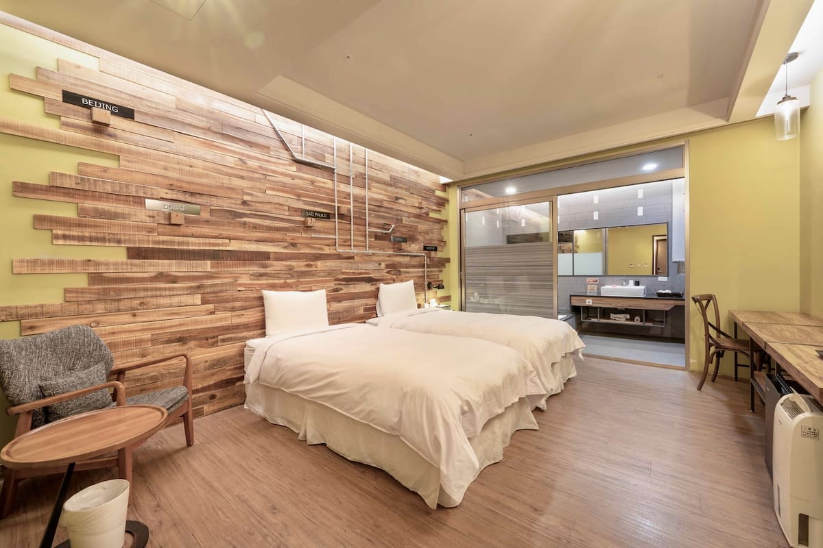A hotel room with a wooden wall, two beds, a chair, a table, and a small kitchenette.
