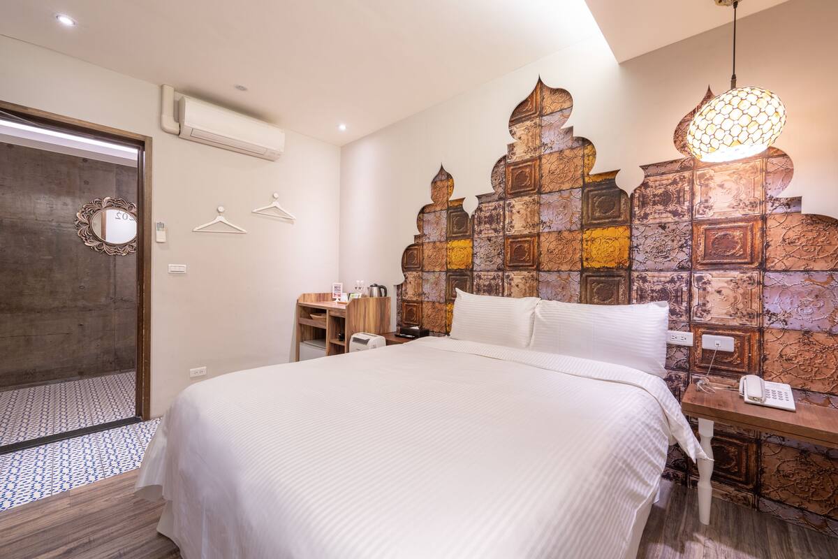 A hotel room with a large bed, a wooden headboard with a unique design, a bedside table with a lamp, and a mirror on the door.