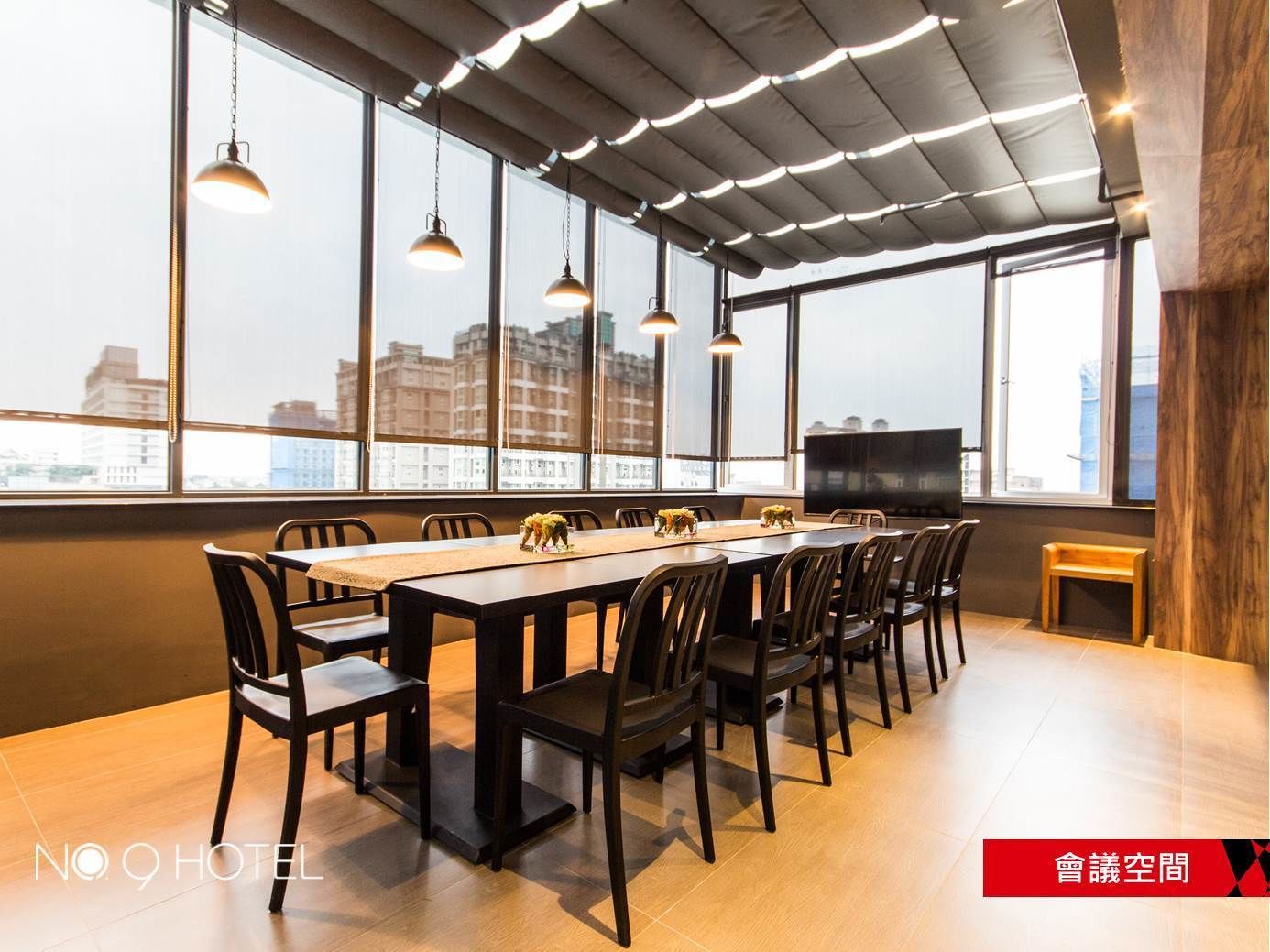 A conference room with a long table, multiple chairs, pendant lights, and large windows offering a city view.