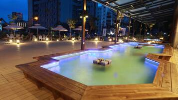 A heated outdoor swimming pool area with wooden benches and umbrellas, surrounded by a covered walkway.