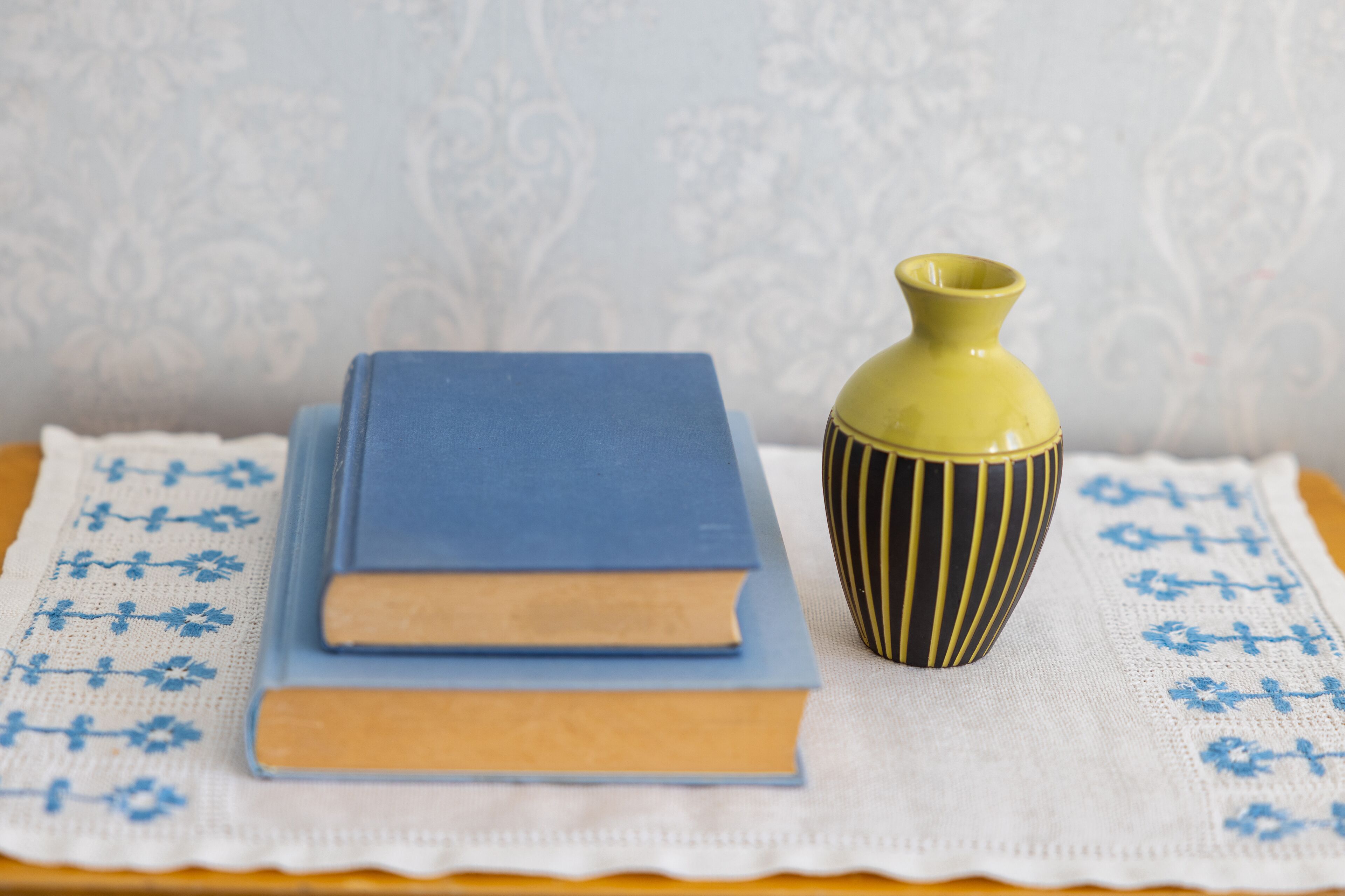 Una mesa con mantel de encaje blanco, dos libros apilados y un florero a rayas.